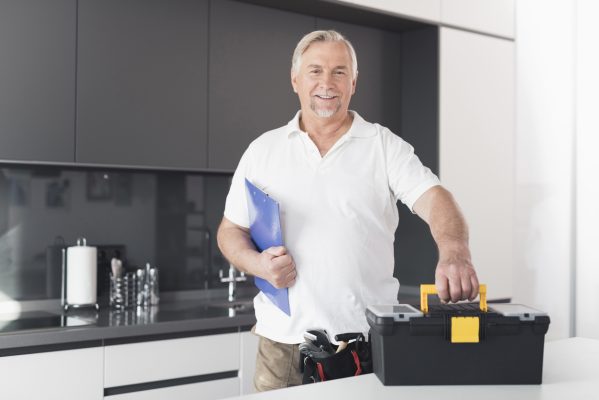 The man is in the kitchen. He holds a form for papers in his hands. Next to him stands a black box for the instrument. A man is in a modern kitchen.